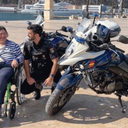 Mujer en silla de ruedas junto a un agente de la Policía Nacional y una motocicleta policial en una zona portuaria.