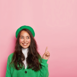 Mujer sonriente con chaqueta y boina verdes que señala hacia arriba con un dedo sobre un fondo rosa.