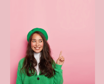 Mujer sonriente con chaqueta y boina verdes que señala hacia arriba con un dedo sobre un fondo rosa.