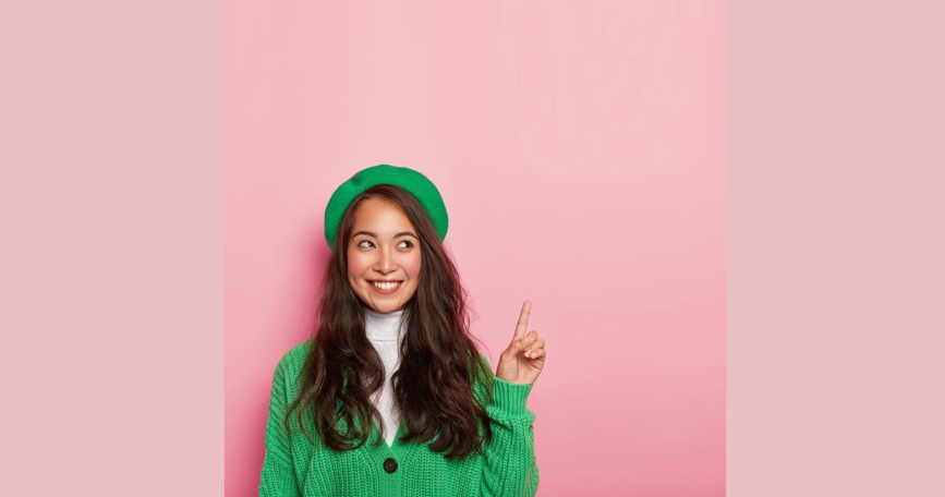 Mujer sonriente con chaqueta y boina verdes que señala hacia arriba con un dedo sobre un fondo rosa.