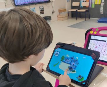Alumno utilizando una tableta educativa junto a un dispositivo de comunicación con pictogramas en un aula.
