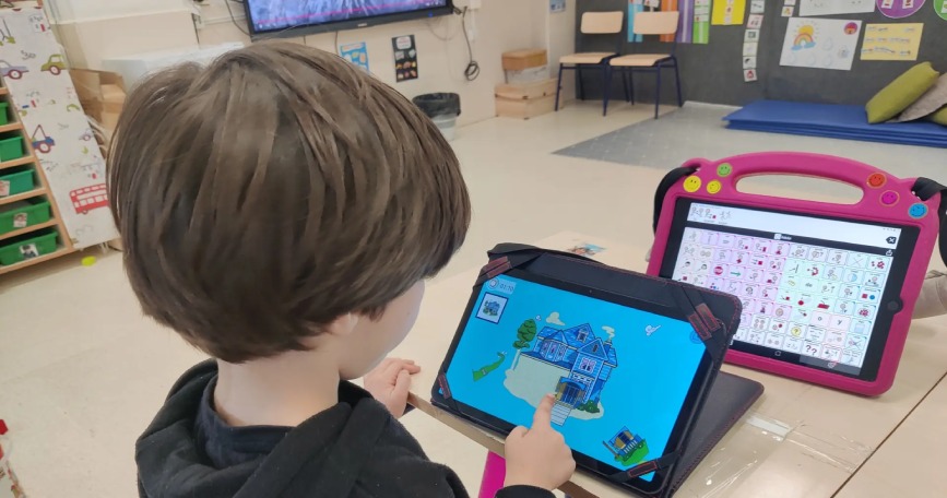 Alumno utilizando una tableta educativa junto a un dispositivo de comunicación con pictogramas en un aula.