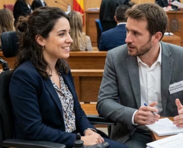 Mujer usuaria de silla de ruedas eléctrica conversa con un hombre junto a una mesa con documentos en una sala de vistas judicial.