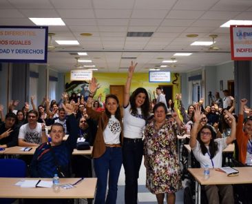 Participantes en una jornada de formación de FAMDIF en Totana Fotografía de grupo de participantes en una jornada de formación, con varias personas levantando los brazos y sonriendo en una sala con mesas y sillas. Algunas utilizan silla de ruedas.