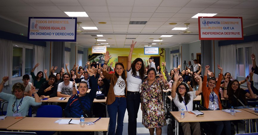 Fotografía de grupo de participantes en una jornada de formación, con varias personas levantando los brazos y sonriendo en una sala con mesas y sillas. Algunas utilizan silla de ruedas.