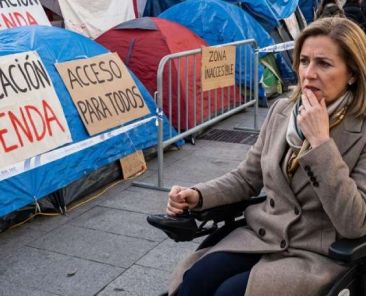 Accesibilidad y movilidad en la acampada de la Puerta del Sol Mujer en silla de ruedas eléctrica junto a una acampada con tiendas, vallas y carteles sobre vivienda y accesibilidad.