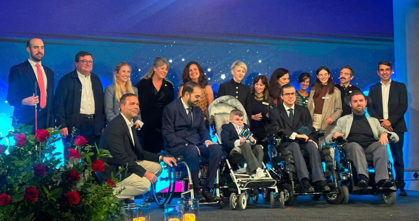 Fotografía de grupo durante una entrega de premios, con varias personas de pie y otras en sillas de ruedas sobre un escenario iluminado en azul.