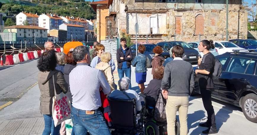 Grupo de personas, algunas en sillas de ruedas, reunidas en una calle de la Ribera de Deusto junto a un antiguo edificio.
