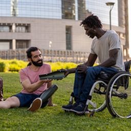 Vida independiente y participación social – Premios FEKOOR Sariak 2026 Dos hombres, uno de ellos usuario de silla de ruedas, comparten una actividad con una tableta electrónica en un parque urbano.