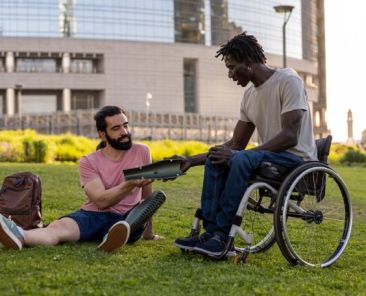 Vida independiente y participación social – Premios FEKOOR Sariak 2026 Dos hombres, uno de ellos usuario de silla de ruedas, comparten una actividad con una tableta electrónica en un parque urbano.
