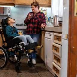 Prestación CUME y cuidados familiares de menores con discapacidad Un niño con discapacidad en silla de ruedas conversa con una mujer adulta en la cocina de su vivienda.