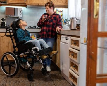 Prestación CUME y cuidados familiares de menores con discapacidad Un niño con discapacidad en silla de ruedas conversa con una mujer adulta en la cocina de su vivienda.