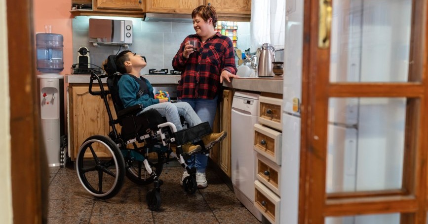 Un niño con discapacidad en silla de ruedas conversa con una mujer adulta en la cocina de su vivienda.